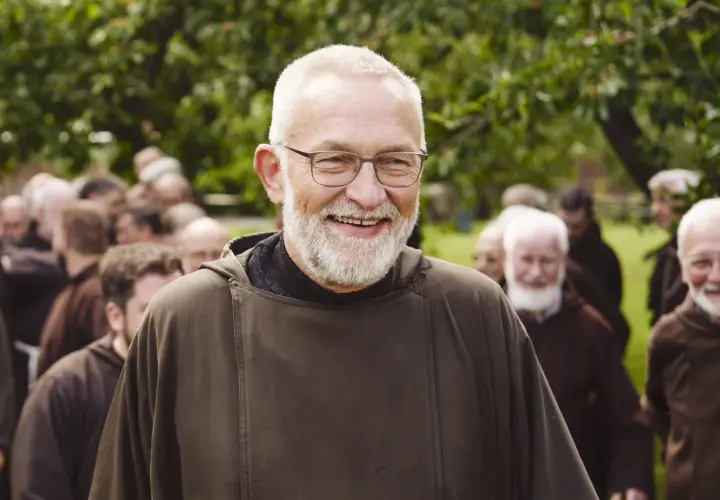 Bruder Paulus, ein älterer Mann mit Brille und Bart, lächelt in die Kamera. Hinter ihm stehen weitere Männer in braunen Kutten.