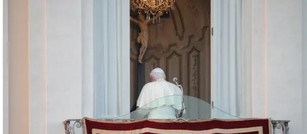 Der Papst steht auf dem Balkon, hinter ihm ein Kruzifix und ein Kronleuchter.